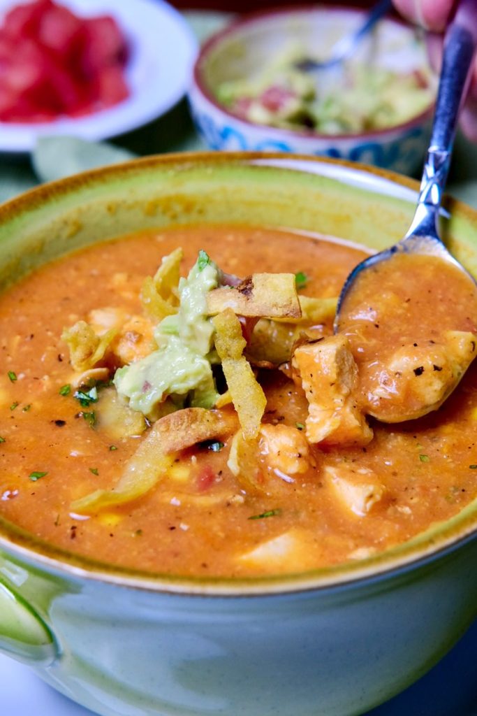 Spoon shot of Creamy Chicken Tortilla Soup garnished with tortilla strips and guacamole.  Condiment bowls of chopped tomato and avocado in background.