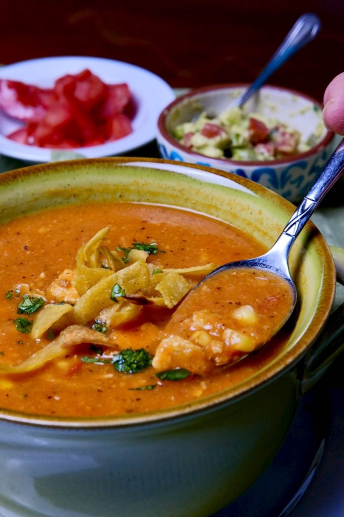 Creamy Chicken Tortillas soup in green soup bowl with spoon shot.  Condiment bowls with chopped tomato and guacamole in background 