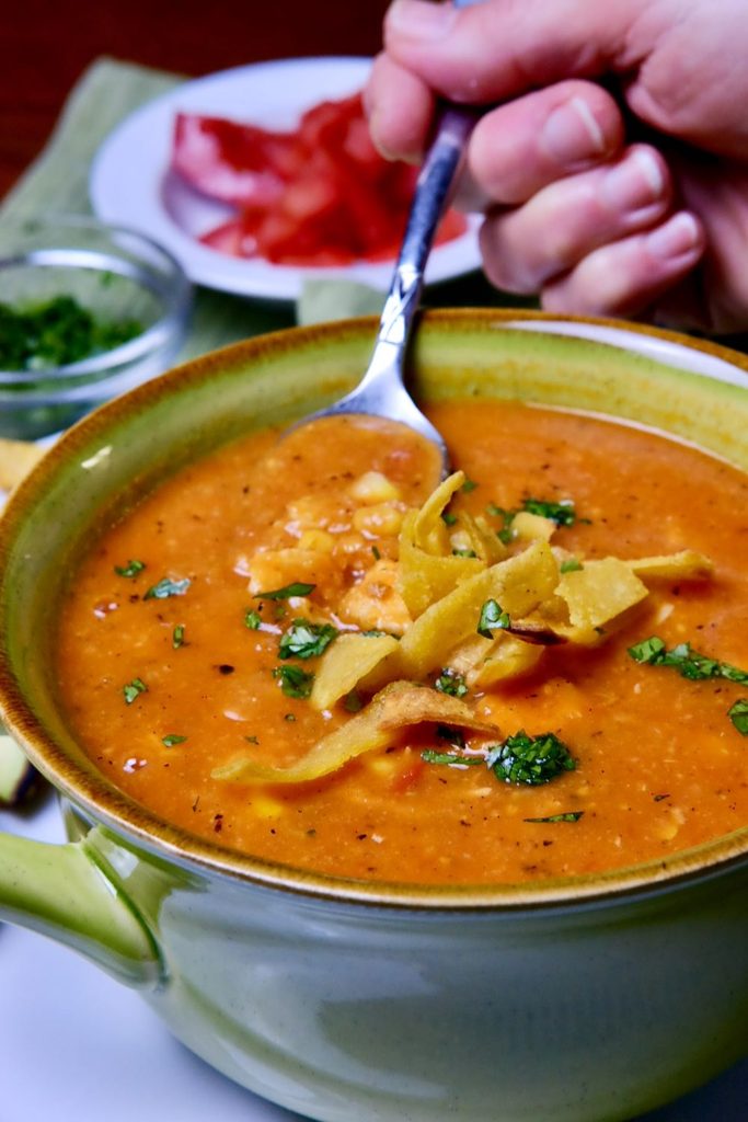 Spoon shot of Creamy Chicken Tortilla Soup in green bowl garnished with homemade tortilla strips and cilantro.  Condiments of chopped tomato, cilantro and avocado in background.