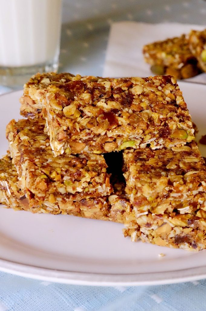 Granola bars stacked on white plate set on blue and white linen with glass of milk in background>
