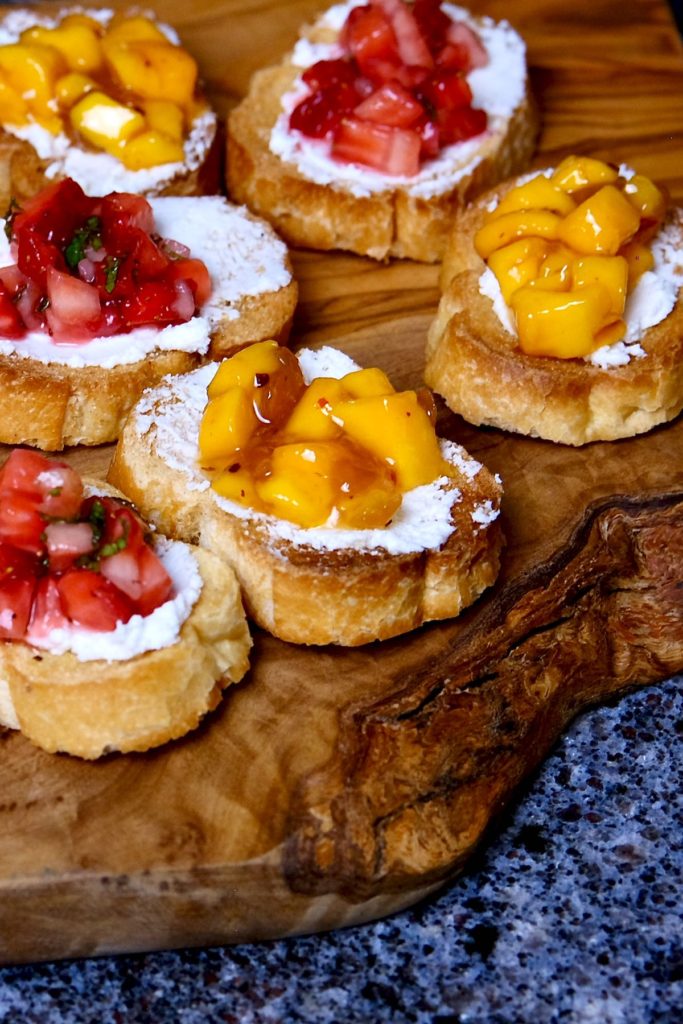 Bourbon mango salsa and strawberry salsa served over bruschetta spread with goat cheese.  Both strawberry and mango appetizers are served on a olive wood board.