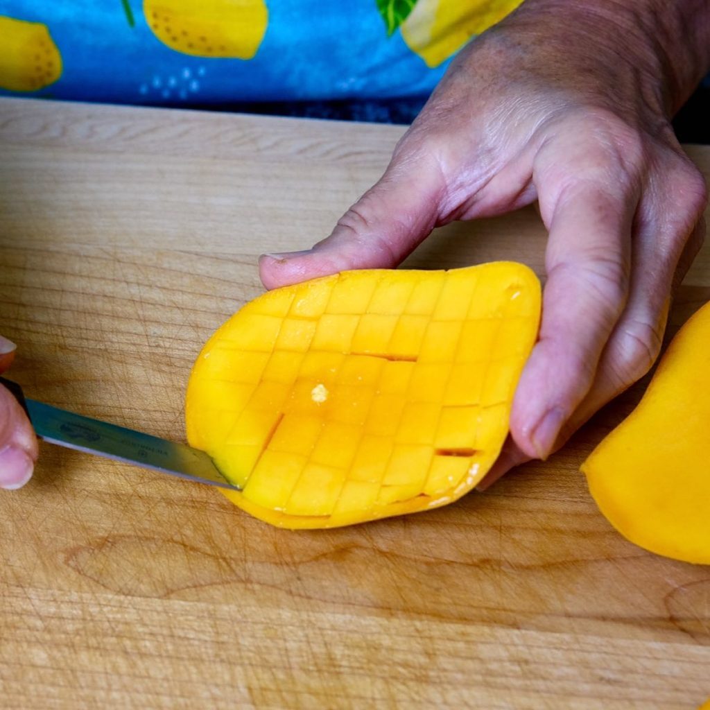 Mango being cut into cubes on maple cutting board with pairing knife.