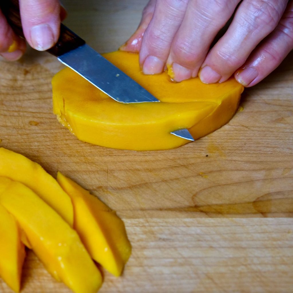 Slicing flesh away from sides of mango pit. Mango slices sitting in foreground on maple cutting board.