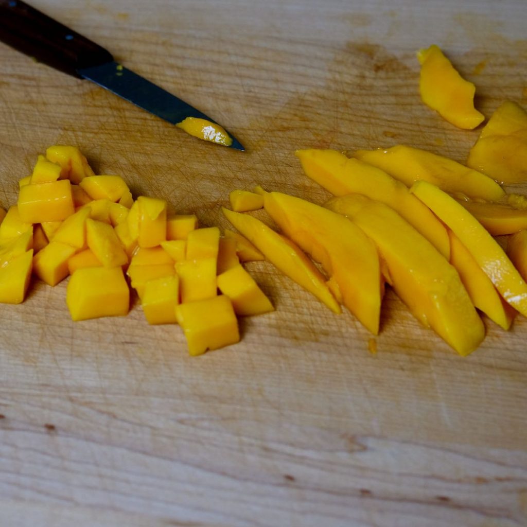 Mango cubes and Mango slices on maple cutting board with knife in background.