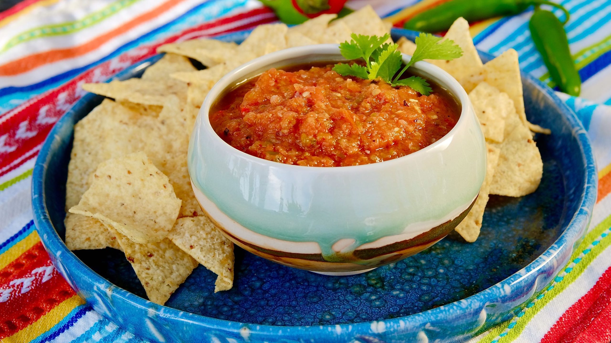 Fire Roasted Salsa in a glazed bowl set on a blue plate surrounded by tortilla chips. Backdrop linen is multi-colored stripes.