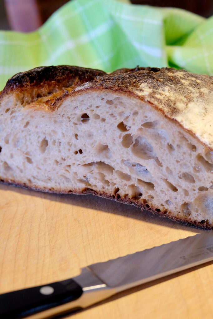 Crusty Artisan Bread loaf sliced open revealing airy crumb texture is set on a maple wood board with lime green linen towel in background and bread knife in foreground>