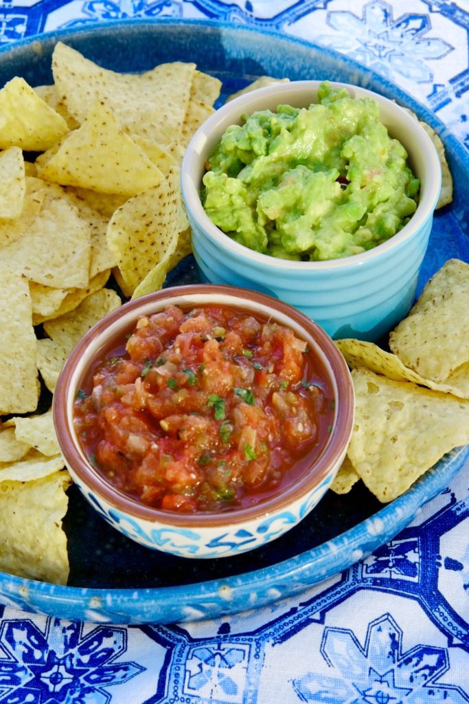 Fire roasted salsa in white and blue bowl set on a blue plate of tortilla chips.. Turquoise bowl of guacamole in background set on blue and white linen.