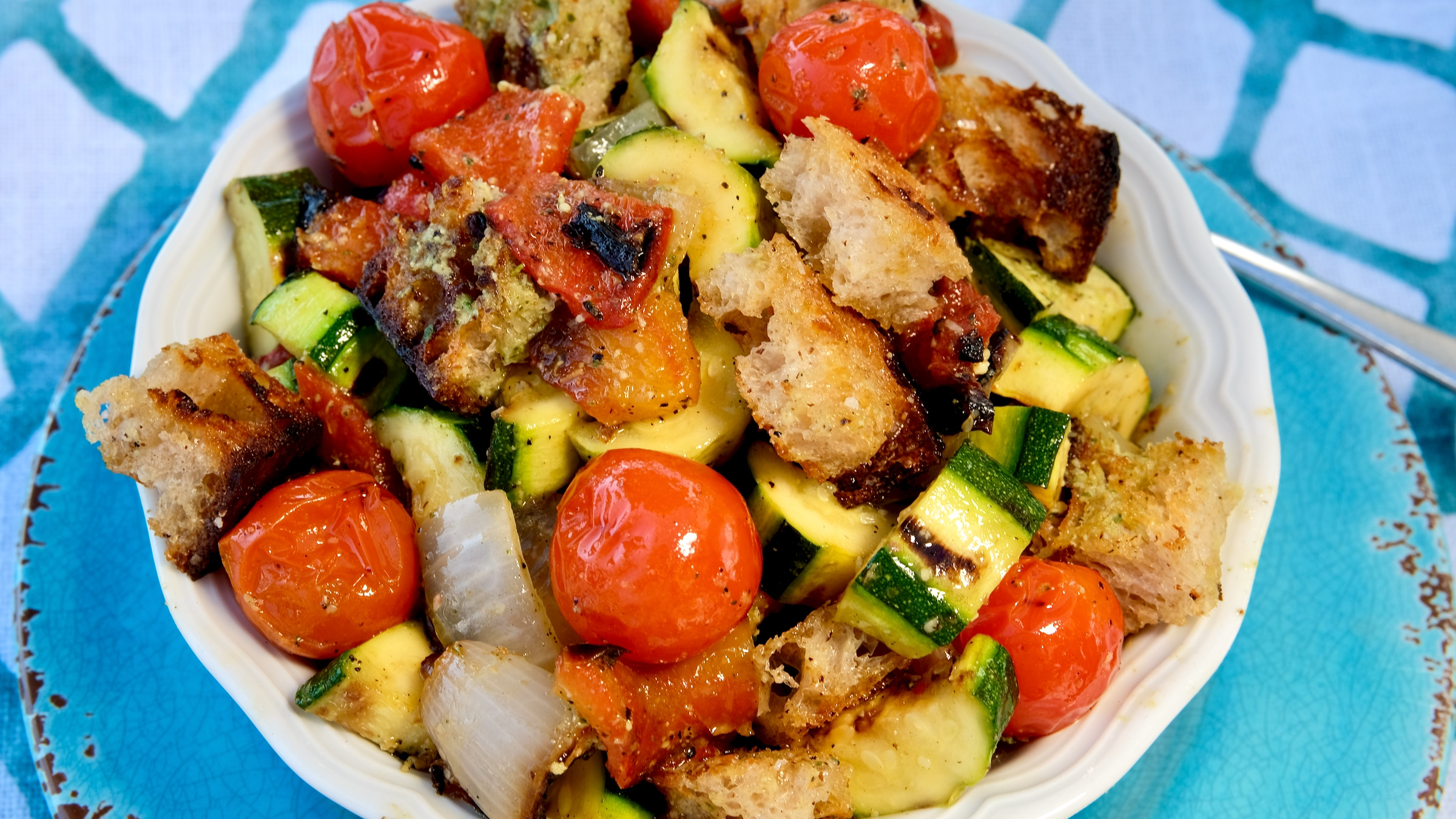 Grilled Vegetables and Bread tossed together with dressing in a white bowl set on a turquoise charger plate. Background is a white and turquoise linen.