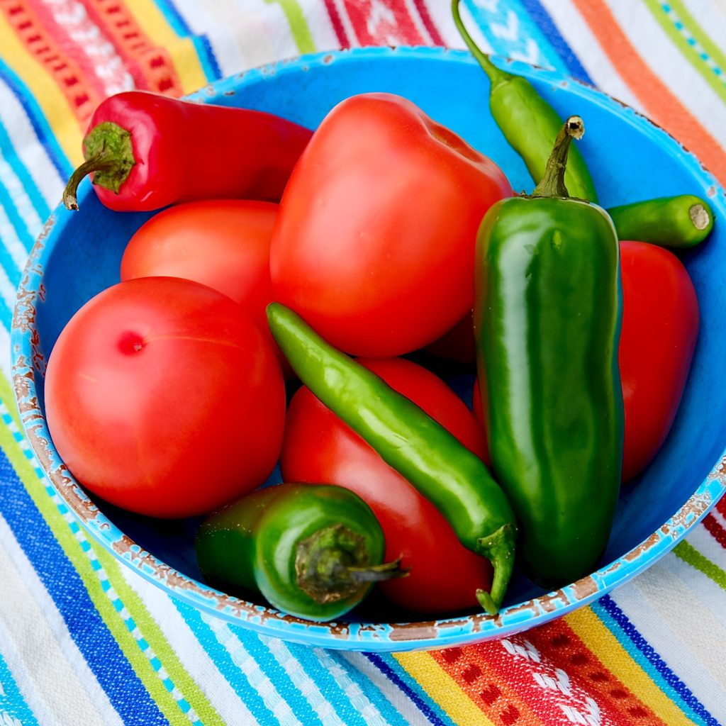 Blue bowl set on multi-colored striped towel filled with fresh roma tomatoes, red and green Jalapenos and green Serrano chilies.