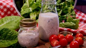 Two bottles of light ranch dressing set on wood board with baby tomatoes, chives, knife and red and white checkered linen in background with bowl of chopped lettuce.