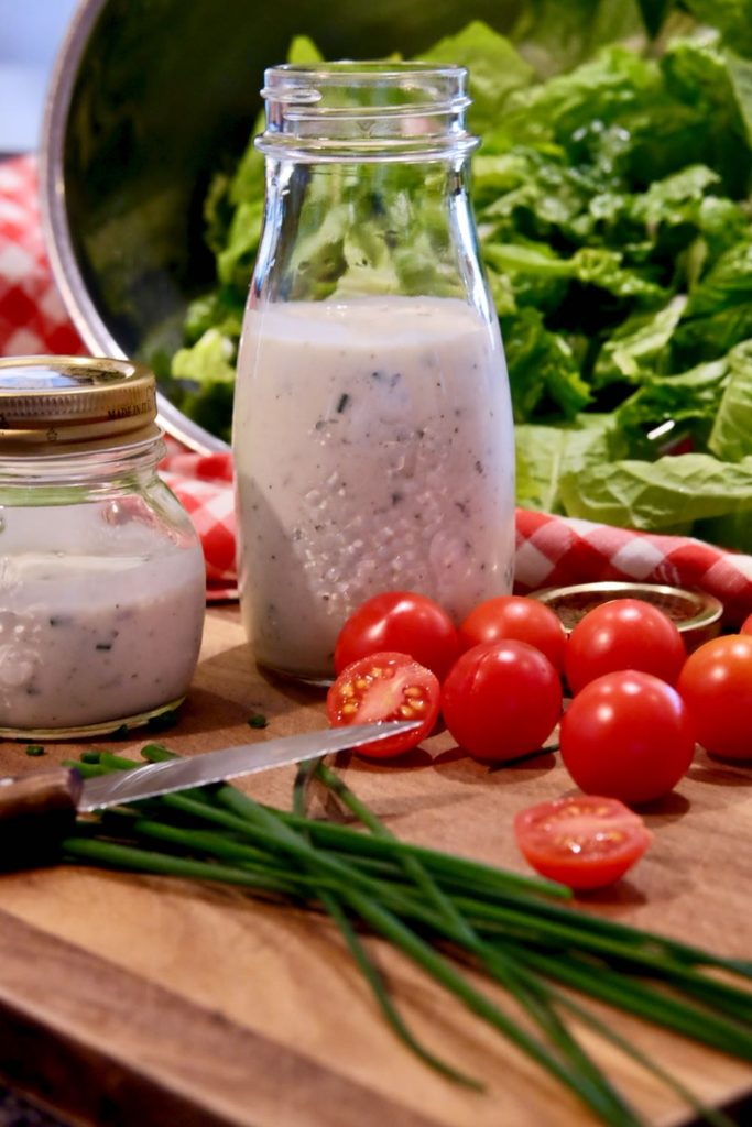 Two bottles of light ranch dressing set on wood board with baby tomatoes, chives, knife and red and white checkered linen in background with bowl of chopped lettuce.