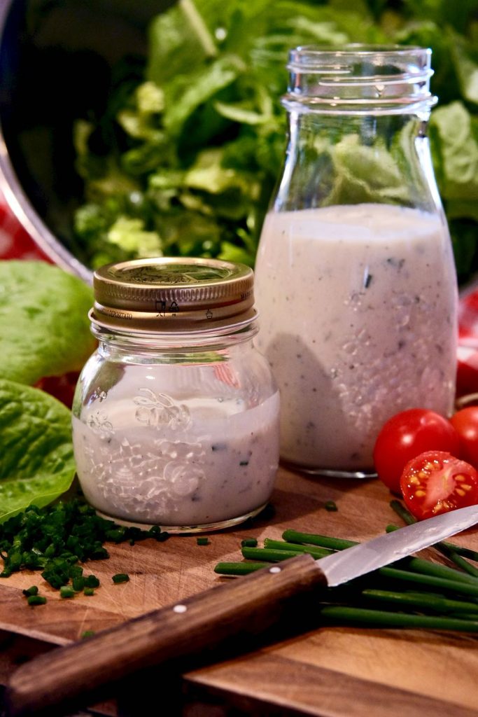 Glass bottles of both Light Ranch and Light Buttermilk Ranch set on a wood board with a bowl of cut lettuce in background and chopped chives and baby tomatoes are in foreground.