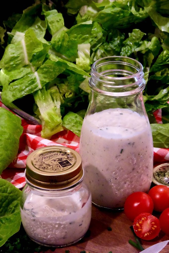 Glass bottles of both Light Ranch and Light Buttermilk Ranch set on a wood board with a bowl of cut lettuce in background and chopped chives and baby tomatoes are in foreground.