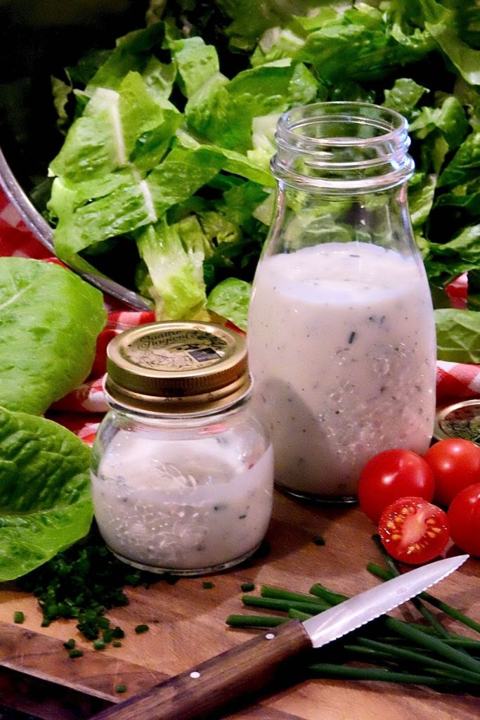 Glass bottles of both Light Ranch and Light Buttermilk Ranch set on a wood board with a bowl of cut lettuce in background and chopped chives and baby tomatoes are in foreground.