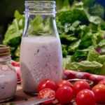 Light Ranch Dressing in a glass bottle set on a walnut board with a bowl of chopped lettuce in background. Baby tomatoes and a knife set in foreground.