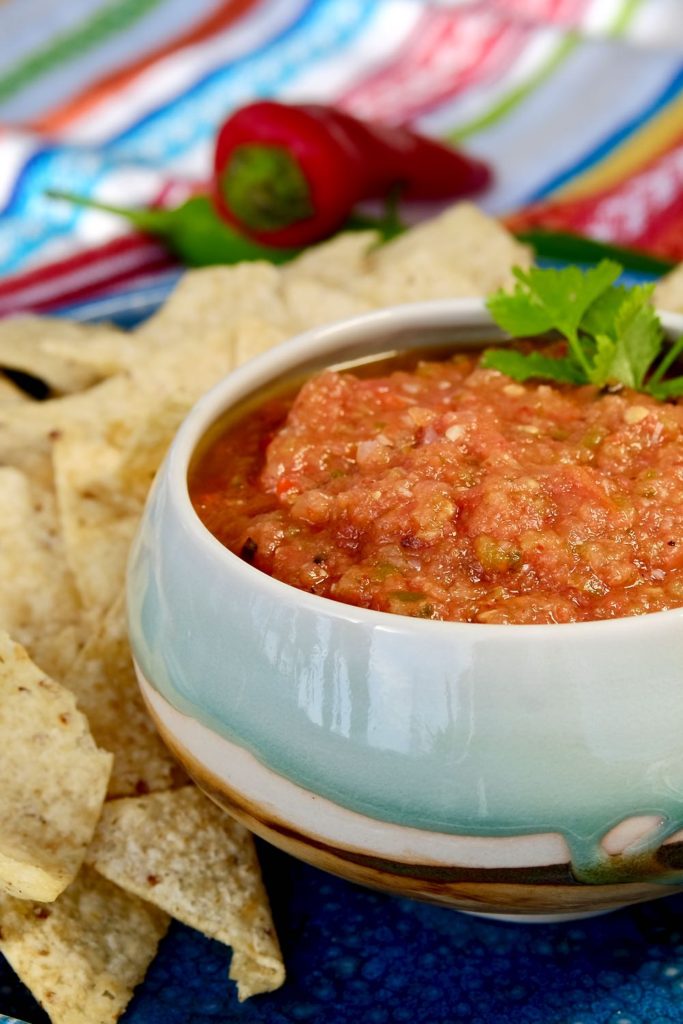 Blue and brown glazed bowl filled with roasted salsa set on a blue plate of tortilla chips.  Multi-colored stirpped linen with red chili pepper in background. 