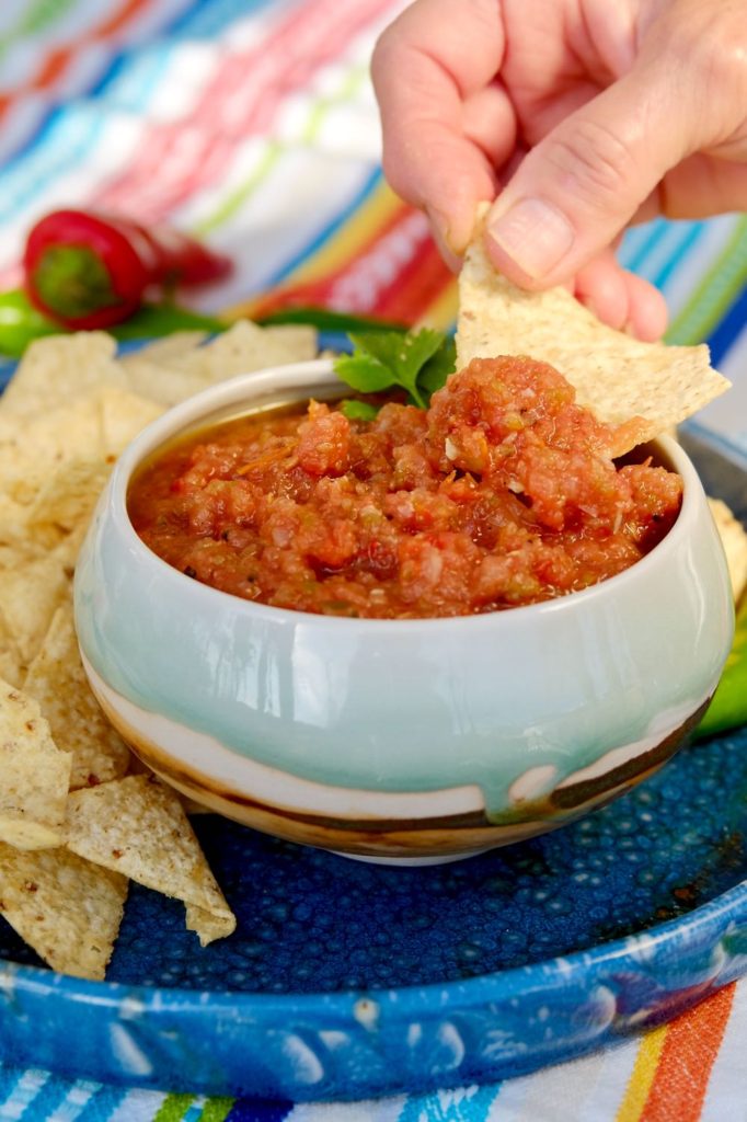 Blue and brown glazed bowl filled with roasted salsa set on a blue plate of tortilla chips. Multi-colored stirpped linen with red chili pepper in background.