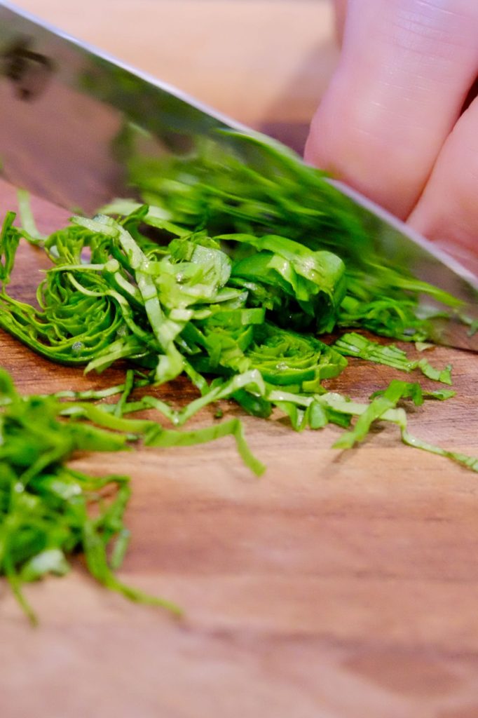 Basil being cut into thin ribbons (chiffonade) on walnut cutting board.