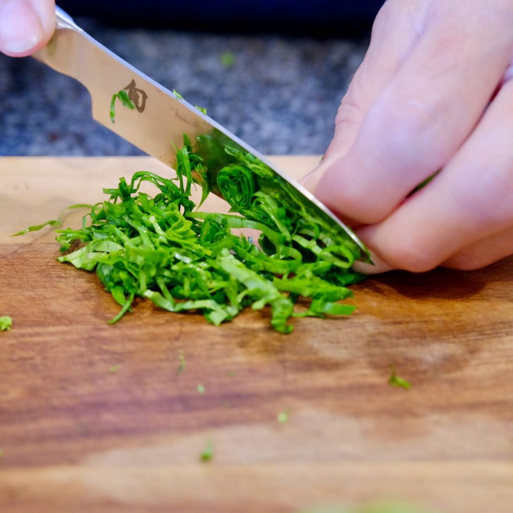 Basil being cut into shredded ribbons for garnish on a walnut board.