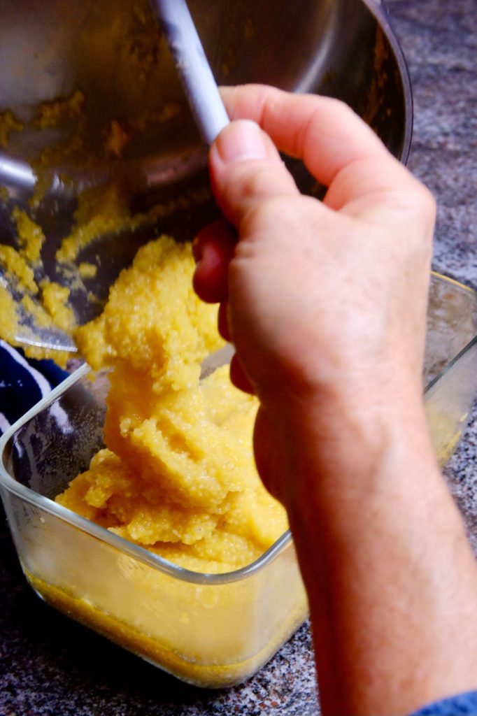 Leftover polenta being spooned out of a stainless pan into an oiled glass dish.