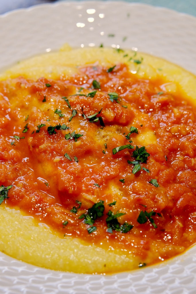 Cooked Polenta served in white rimmed bowl topped with a marinara sauce garnished with chopped fresh parsley.