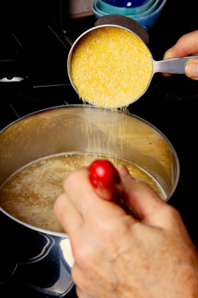 Grains of polenta slowly being stirred into boiling pot of broth set on a stove.