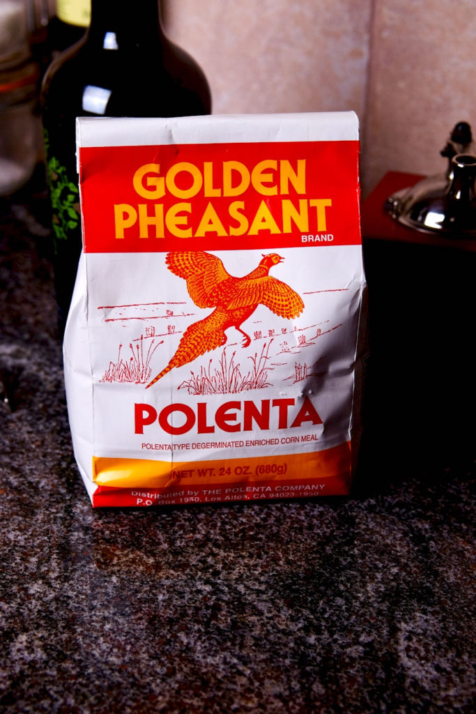 A bag of Golden Pheasant Polenta is set on black granite counter with olive oil bottle in background.  Polenta package is a white bag with red and yellow lettering and images.