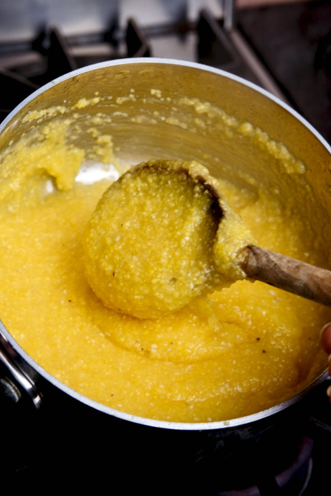 Basic polenta in final stage of cooking in stainless steel pan. Olive wood spoon held up showing desired thickness of finished polenta. 