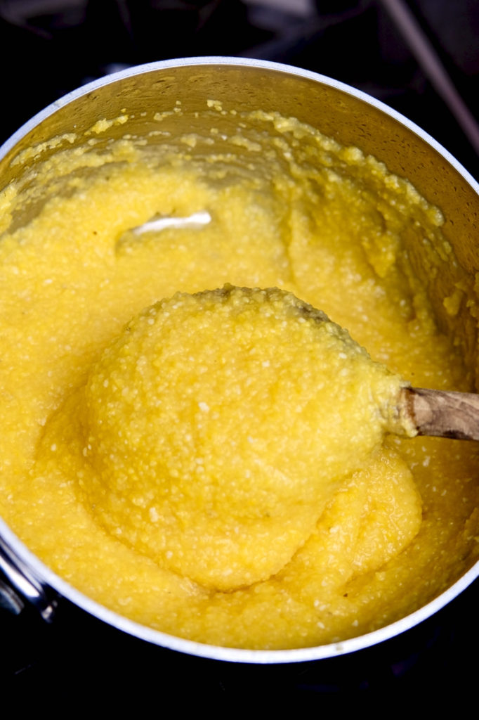 Basic polenta in final stage of cooking in stainless steel pan. Olive wood spoon held up showing desired thickness of finished polenta.