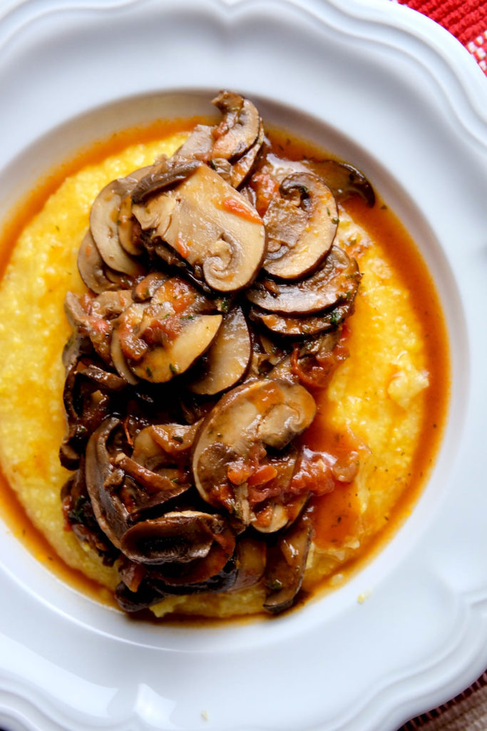 Basic polenta served in a white rimmed bowl is topped with Mushroom Tomato Ragu and garnished with fresh minced parsley.