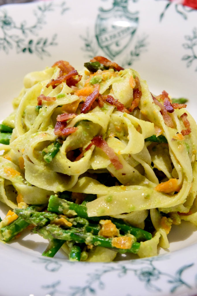 Pasta Primavera with carrot ribbons and asparagus tossed with Pea and Pistachio Pesto and garnished with bacon bits served in a white and green Italian pasta bowl.