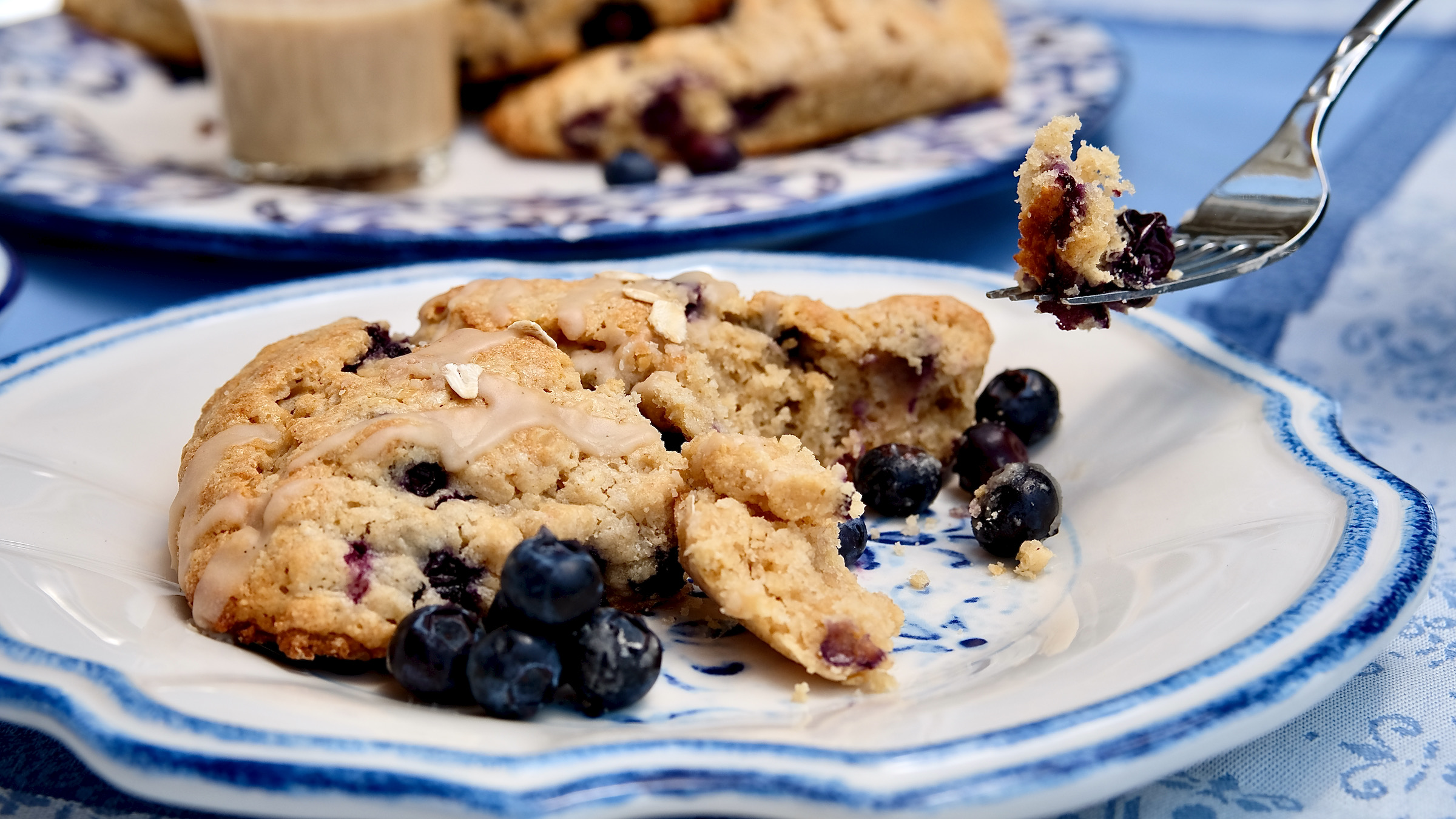 Oatmeal and Blueberry Scone served on a white and blue plate showing a bite shot of scone. Blue and white platter of scones with maple glaze is set in the background.