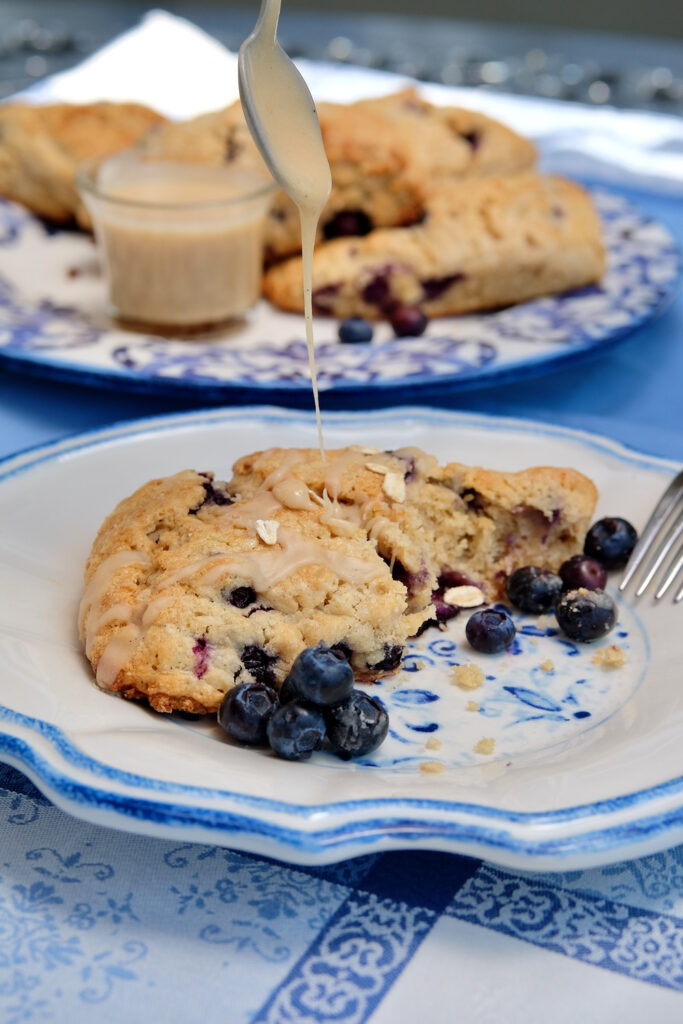Oatmeal and blueberry scones on a blue and white plate being drizzled with maple glaze.  Plate is set on blue and white linen.  Additional scones on blue and white serving plate with glass vessel of maple glaze.