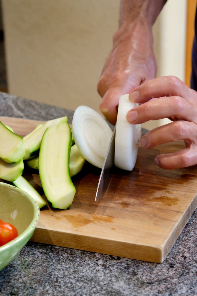 Onion being cut into desired 3/4 inch thickness on a walnut cutting board.  Zucchini halves are piled on the left side of board with a bowl of cherry tomatoes in foreground of left corner.