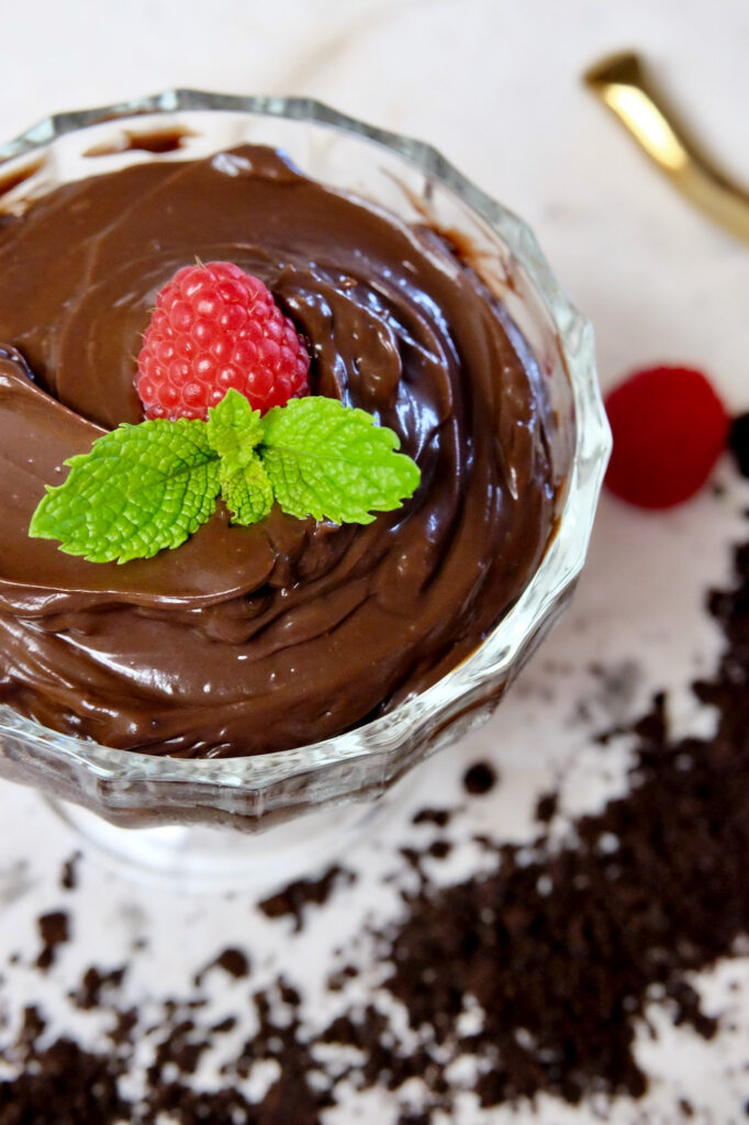 Overhead shot of chocolate pudding in a glass dessert bowl garnished with fresh mint and raspberry is set on a white marble background.  Chocolate sand is scattered around base of glass with fresh raspberries. 