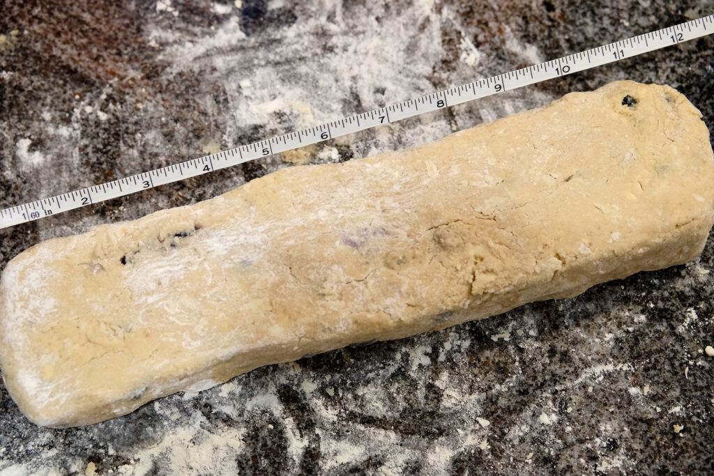 Formed dough log on floured marble surface with tape measure paralleled to dough depicting 12 inch length.