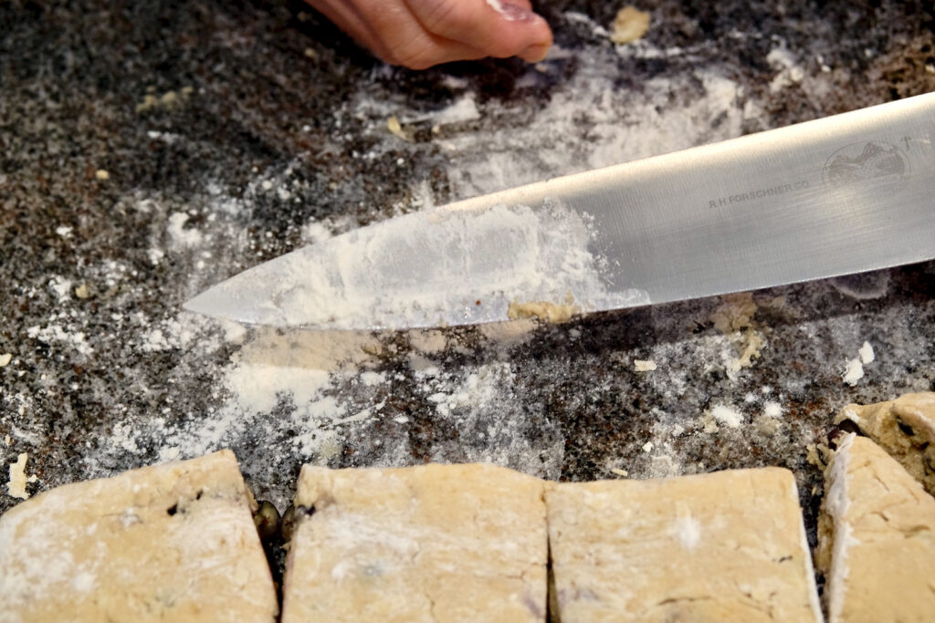 Picture shows knife held over marble surface being dusted with flour in between each cut.  Four cut squares of dough are in forefront.