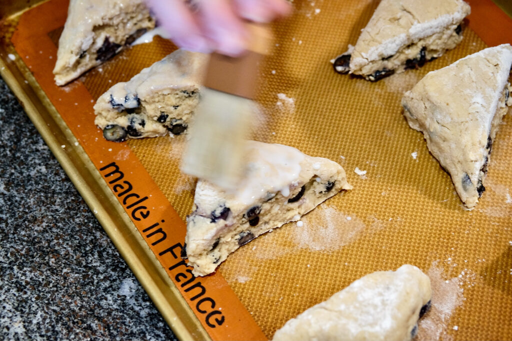 Scone wedges placed on baking sheet lined with Silpat being brushed with cream.