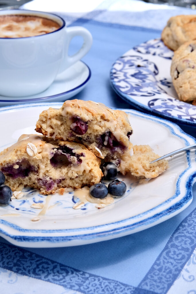 Oatmeal and Blueberry Scone split in half served on a white and blue plate and drizzled with maple glaze.  Blue and white serving plate with scones and coffee cup is in background set on blue linen.