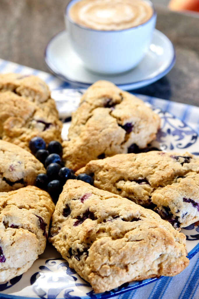 Oatmeal Blueberry Scones are served on a blue and white platter and garnished with a cluster of fresh blueberries in center of plate.  A cafe latte in a blue and white cup and saucer is in background
