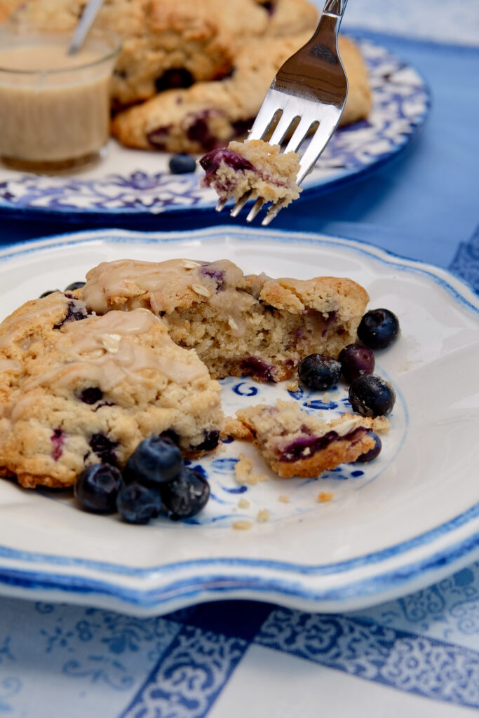 Blueberry Oatmeal Scone broken open on a white and blue rimmed plate depicting bite shot on fork above scone.   Serving plater of scones with glass carafe of maple glaze is in background set on blue linen.