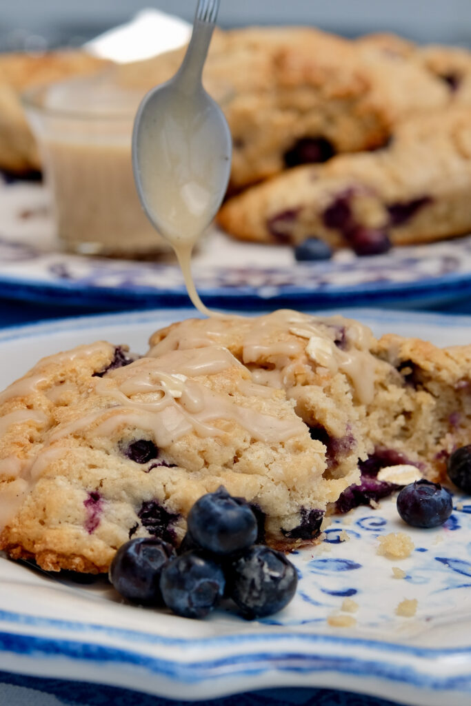 Oatmeal Blueberry Scone served on a blue and white rimmed plate and garnished with fresh blueberries.  Spoon hovering above scone as maple glaze is drizzled onto scone.  Serving plate with additional scones and glass carafe of maple glaze is in background.