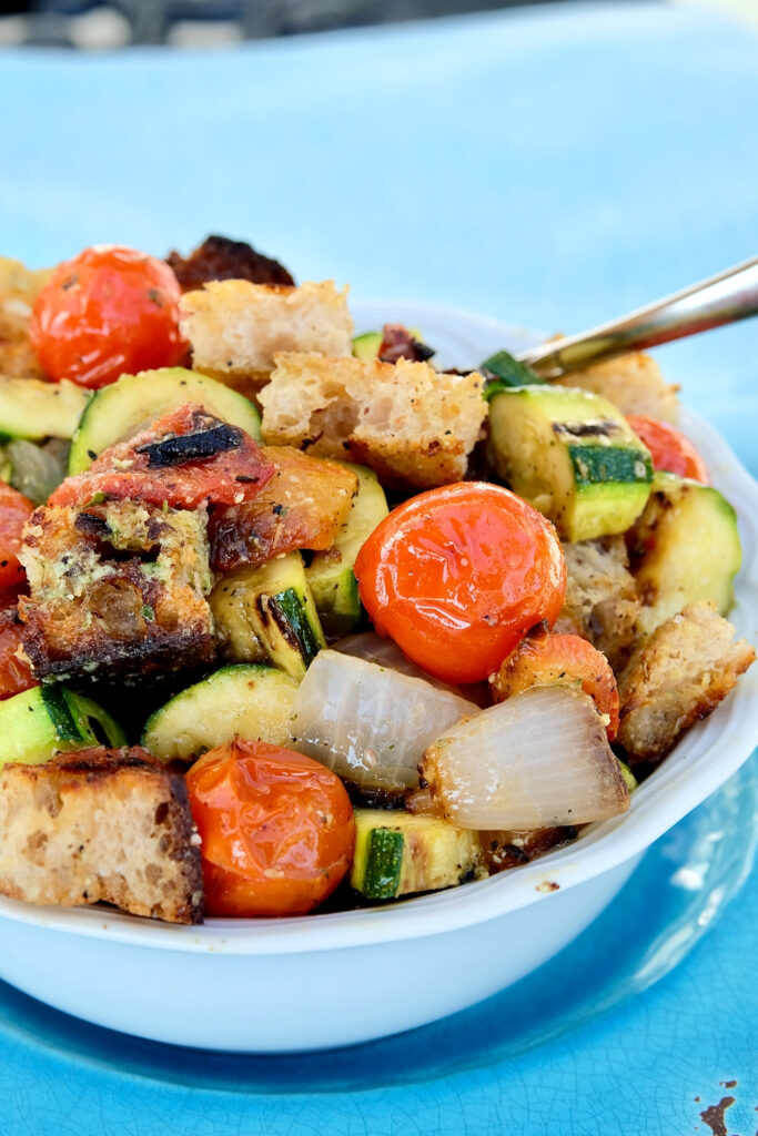 Grilled tomatoes, onion, zucchini and bell pepper tossed together with cubed grilled bread and dressing served in a white bowl set on a blue plate.  Serving spoon is setting in bowl in back.  