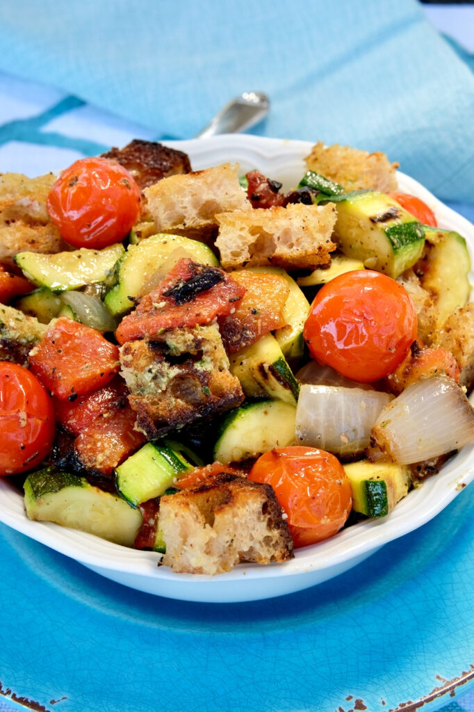 Grilled vegetable panzanella salad is tossed with dressing and served in a white bowl set on a turquoise charger.  Blue and white linen are in background with serving spoon.