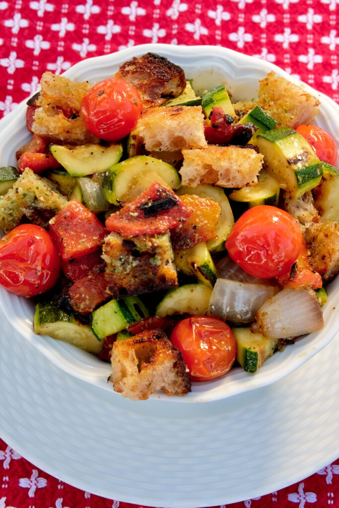 Panzanella Salad of grilled vegetables and bread tossed with dressing are served in a white bowl set on a white charger.  Background is red linen with white squiggles. 
