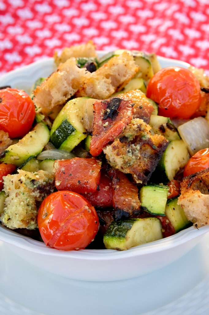 Grilled zucchini, cherry tomatoes, onion and bell peppers tossed with grilled bread cubes and dressing served in a white bowl set on a white plate.  Red linen cloth is in background.