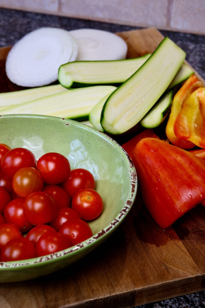 Green bowl of washed cherry tomatoes set on a walnut cutting board next to quartered bell peppers.  Zucchini halves and sliced onion are on board set behind bowl of tomatoes.