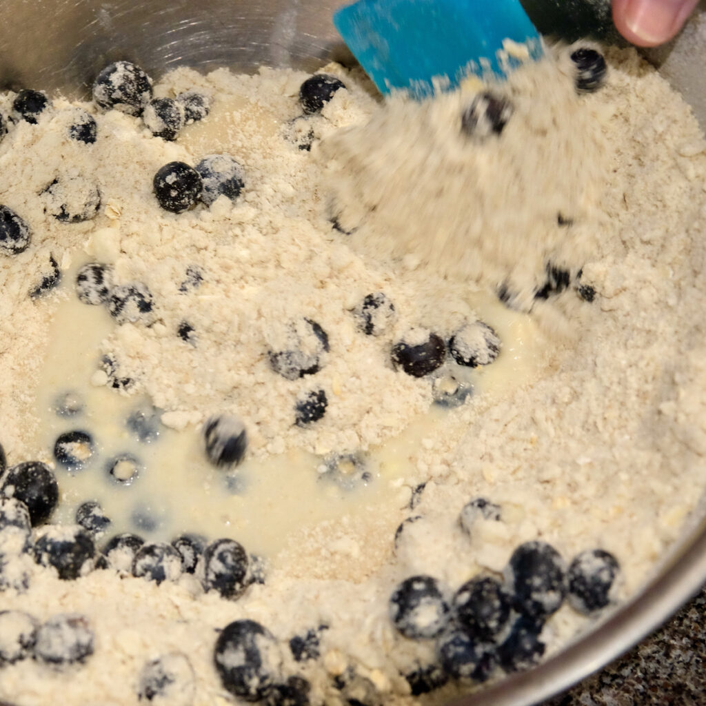 Blue spatula shows pulling dry ingredients from sides of stainless bowl into the center.