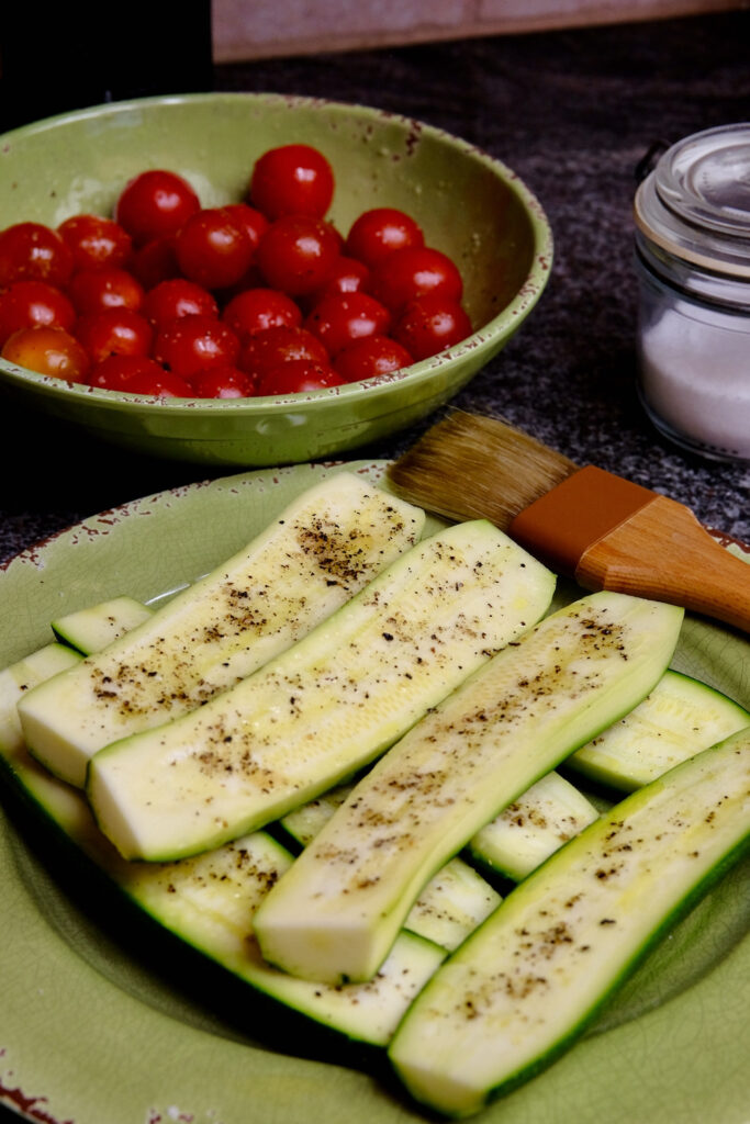 Zucchini halves stacked on a green plate have been brushed with olive oil and seasoned with salt and pepper.  Basting brush is set on upper right corner of plate with a green bowl of seasoned cherry tomatoes in left background and glass container of salt in right corner of background.