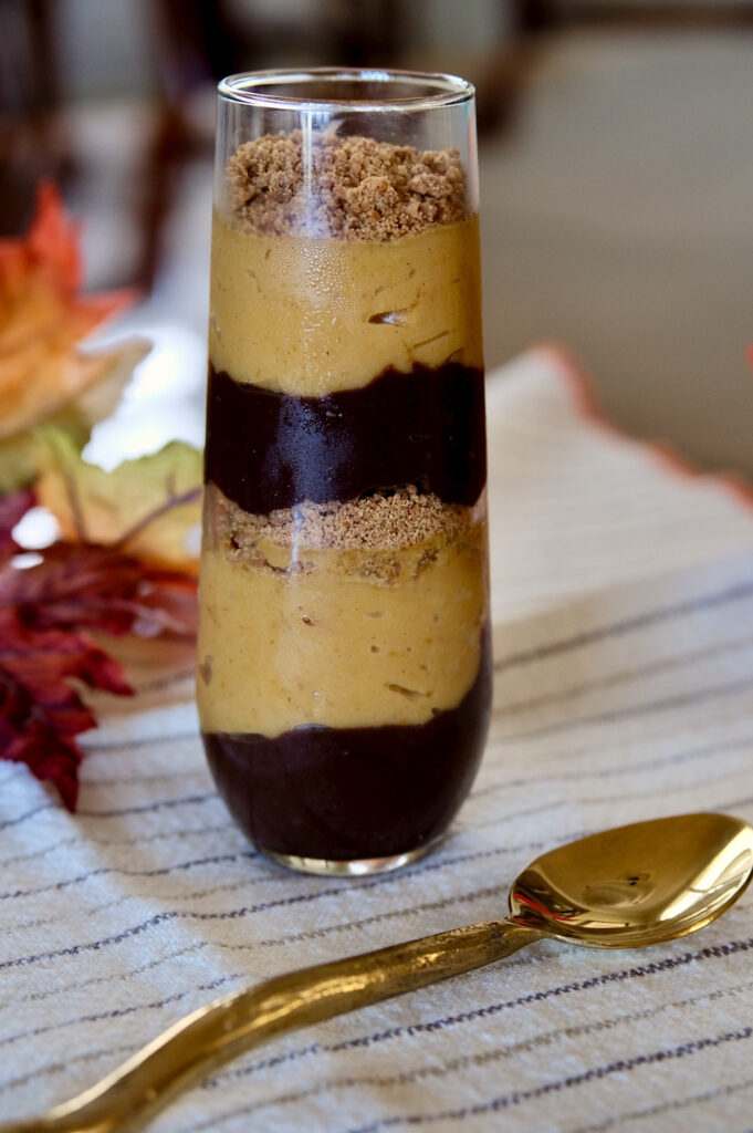 Tall parfait glass with alternating layers of chocolate and pumpkin pudding with edible sand.  Glass is set on a cream colored linen with brown pin stripes.  Gold spoon is resting in foreground and autumn colored leaves are in background>